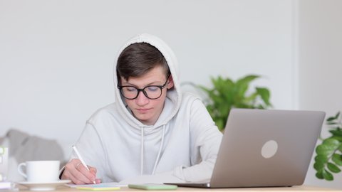 Remote education of teen scholars. Concentrated schoolboy in white hoodie spbi and glasses writes notes in pad near modern laptop at table in living room 4k video