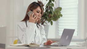 Doctor telehealth remote service. Careful Asian woman general practitioner talks spbi on mobile phone and types message on contemporary laptop in clinic 4k video