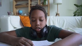 Student studying in front of device screen and using headphones, sitting at table in home room spbd. American African child looks ahead with smile and sits at desk with notebook, puts on headphones