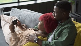 Young married people using smartphone for entertainment and lying on couch in home room spbd. Closeup view of African American woman, man hold phone in hand and look at screen with smiles, watch video