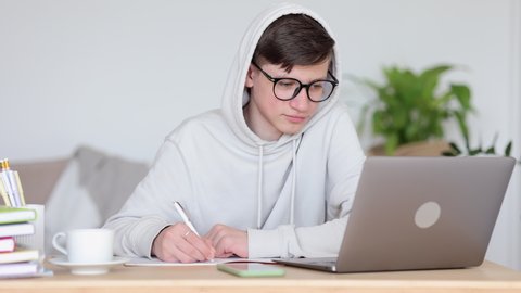 Online home school for teenage students. Serious teen boy in white spbi hoodie does homework at video lesson via laptop at workplace in living room 4k video