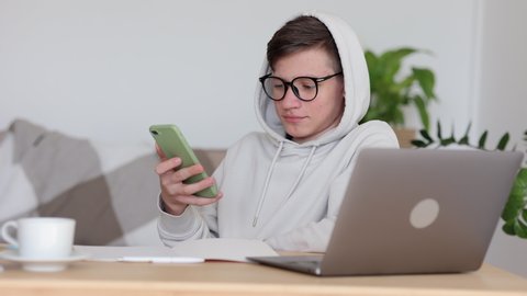 Teenage boy home online schooling. Kid in white hoodie talks on spbi mobile phone using loudspeaking mode near laptop computer at table in living room 4k video