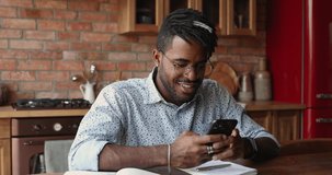 Handsome African hipster guy in glasses and dreadlocks hairstyle sit in kitchen holds smartphone chatting, watch received video, listen favourite band music on-line. Fun, leisure, modern tech concept