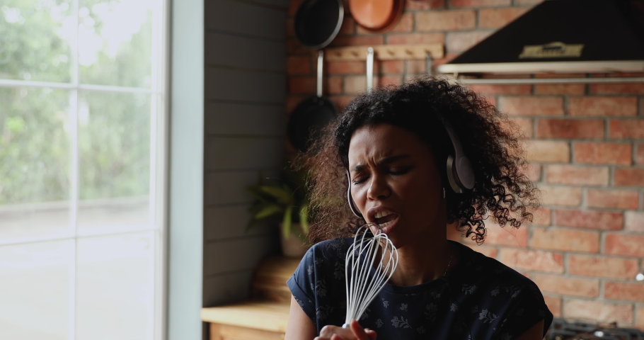 Close up African woman distracted from cooking in kitchen listening tune through wireless headphones sing a song into whisk enjoy hobby. Carefree female passionate in music, karaoke, pastime concept Royalty-Free Stock Footage #1082335528