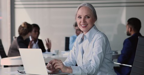 Older businesslady sit at desk with laptop, smile look at camera work on device in office, diverse colleagues on background. Leader, founder, executive manager, corporate staff member portrait concept