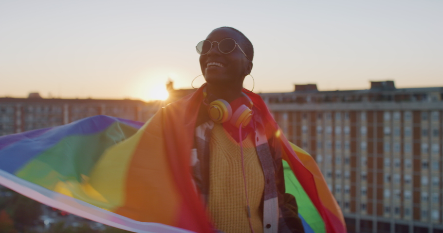 Cinematic shot of young stylish african woman carrying flag of LGBT rainbow symbol and feeling free on urban scape background at sunset. Concept of diversity, equity, peace and love, freedom, liberty.