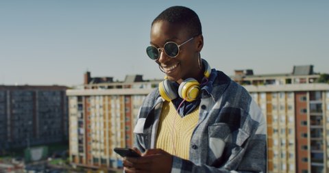 Cinematic shot of young stylish african woman with sunglasses is using smartphone on urban scape background with sun shining. Concept of urban, technology, connection, communication, lifestyle, youth.