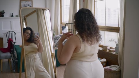 Curly haired overweight young woman blogger wearing comfortable clothes in glasses makes photo posing in front of large mirror in room backside view