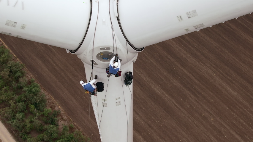 Skilled technician fitters with safety harnesses go down wind turbine pylon blade using long ropes above agricultural plowed field aerial view | Shutterstock HD Video #1082338660