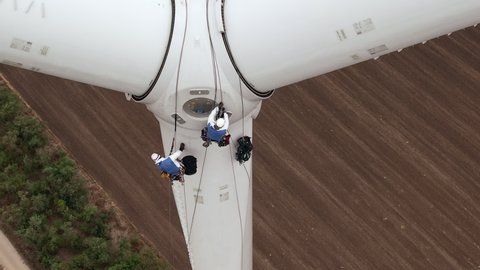 Skilled technician fitters with safety harnesses go down wind turbine pylon blade using long ropes above agricultural plowed field aerial view