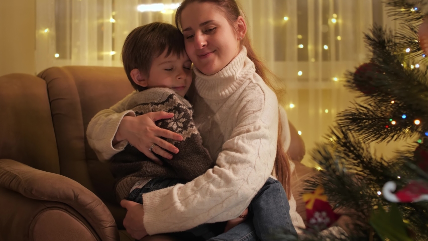 Smiling young mother hugging her son sitting in armchair next to Christmas tree in living room. Families and children celebrating winter holidays