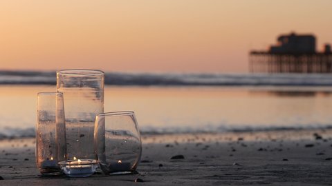 Candle flame lights in glass, romantic beach date by California ocean waves, summer sea water. Candlelight. Glass on littoral sand. Cozy lounge, sunset sky. Wooden pier. Seamless looped cinemagraph