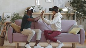 Young multiethnic man and woman in VR headsets sitting on sofa at home, smiling and touching hands of each other while experiencing virtual reality