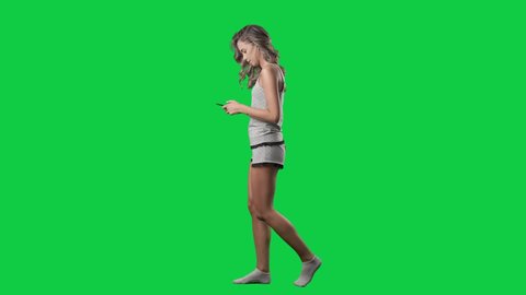 Happy young woman in pajamas walking and using cellphone. Side view. Full body isolated on green screen background