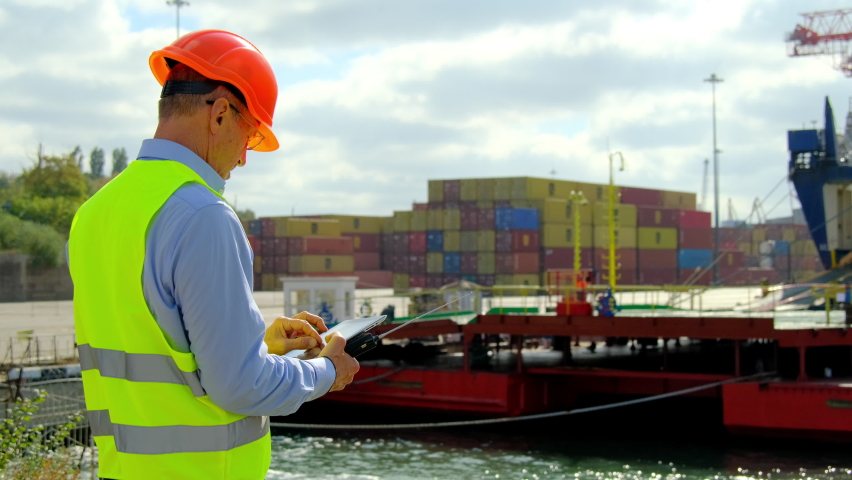 Ship cargo loading process. Safety inspector uniform and helmet marks notes on tablet while worker walk along ferry ramp in dockyard on windy day | Shutterstock HD Video #1082350507