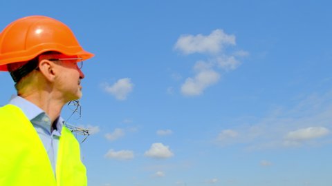 High voltage power lines transmit electric energy under blue sky. Technician in orange helmet looks at built station standing against electric transmission tower