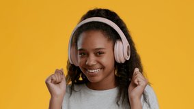 Cheerful Black Teen Girl Listening To Music In Wireless Headphones And Dancing Having Fun Posing Over Yellow Background, Smiling To Camera. Slow Motion, Studio Shot