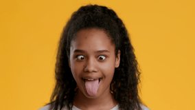 Cheerful African American Teenager Girl Grimacing Sticking Out Tongue Posing And Having Fun Over Yellow Studio Background, Looking At Camera. Headshot Of Female Teen Making Funny Face.