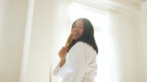 Happy mood concept. Webcam pov portrait of young optimistic asian lady wearing bathrobe singing to camera with hair brush, posing against sun light, slow motion