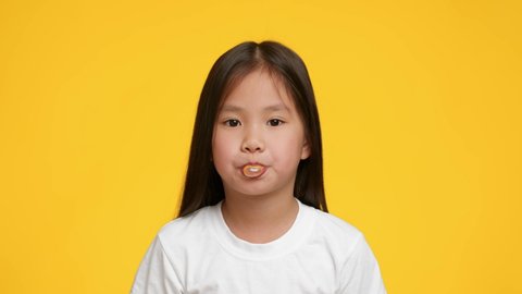 Little Korean Girl Chewing And Blowing Bubble-Gum Posing Standing Over Yellow Studio Background. Unhealthy Nutrition Habits, Junk Food Concept. Slow Motion