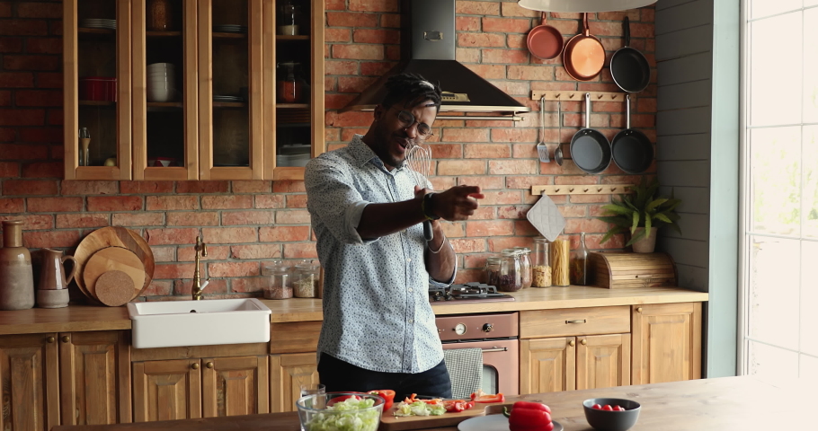 Active African guy holds beater like microphone singing song distracted from preparing vegetarian dinner in kitchen, dancing to energetic pop music, enjoying cooking at home. Karaoke, hobby concept