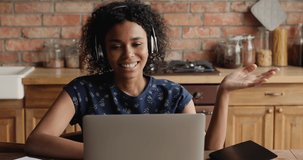 Attractive African woman wear headphones make video call on laptop sit at table in kitchen. Communication through videoconference by personal or business event, online counselling, modern tech concept