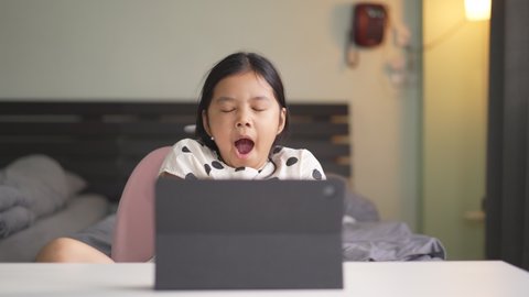 Asian child student learning on computer and sleepy yawn or kid girl studying online class by video call and twisting stretch oneself or person learn from home on back to school by tablet in bedroom