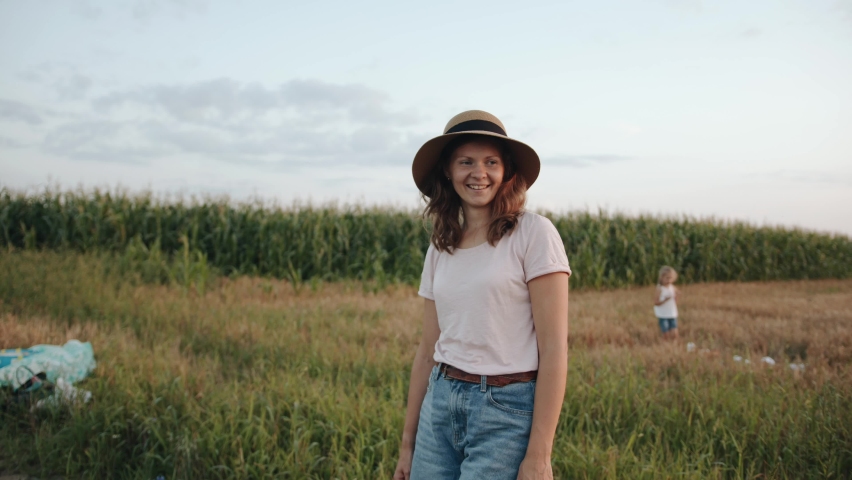 A pleasant young woman stands on the road near a cornfield and adjusts her hat on her head. Slow motion