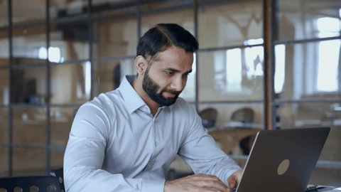 Indian ethnic business man employee professional manager using computer, typing, analyzing digital data working in modern office doing online data market analysis planning looking at laptop.