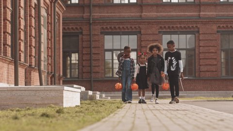 Slowmo shot of four cute African-American kids wearing Halloween costumes trick or treating outdoors running along street with pumpkin buckets holding hands