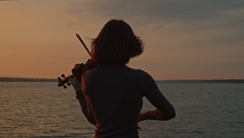 Silhouette of young lady musician playing violin on lakeshore, standing in counter sunrise light, hair is fluttering in wind, Foreground, Slow motion. Royalty-Free Stock Footage #1082364028