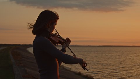 Silhouette of young lady musician playing violin on lakeshore, standing in counter sunrise light, hair is fluttering in wind, Foreground, Slow motion.