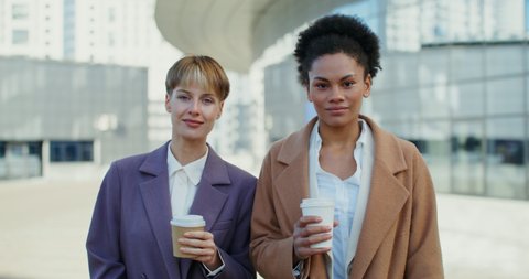 Two business women of different nationalities with glasses with coffee in their hands are smiling looking at the camera, standing outdoors, close-up