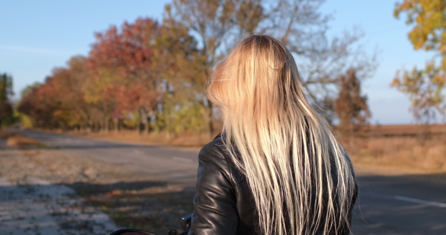 A blonde biker puts a round helmet on her head and takes the wheel of a motorcycle, prepares to move on the road, a beautiful golden autumn. Royalty-Free Stock Footage #1082370613