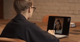 Two young women talking, video call using laptop on wooden desk in a loft coworking. Woman in eyeglasses gesturing, online conversation. Concept of remote meeting