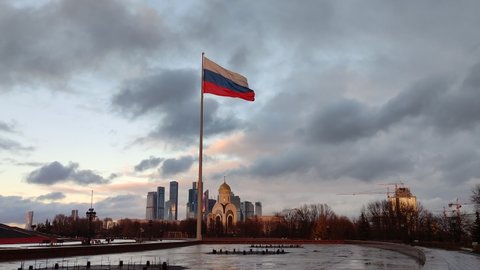 Moscow, Russia - November 13, 2021: Enormous Russian flag waving during clear sky daytime on a flagpole in front of beautiful church and city skyline.