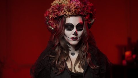 A brunette woman in a beautiful Calavera-style image on All Saints' Day performs a ritual in a dark room with red light from a campfire. Halloween makeup with ram horns, flowers, sewn mouth