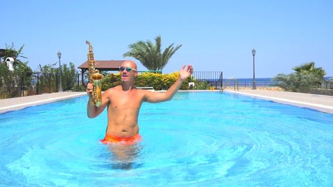 Blurred defocused male musician saxophonist with naked torso in orange swimming trunks dancing, having fun in pool, playing golden alt saxophone on musical instrument. Summer party. Slow motion video.