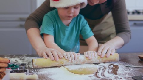 little baby boy child rolling out flatten dough with mother helping. making Christmas gingerbread cookies together in kitchen. 3-4 year kid caucasian in red Santa hat and green t-shirt learning cook