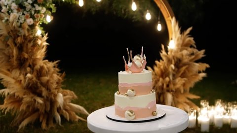 The garden is decorated for a wedding party at night, with a wedding cake in the center. White and pink wedding cake with creative decor. Close up shot