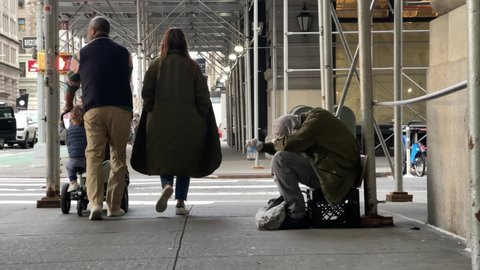 NYC, USA - NOV 8, 2021: family stroller people walking past homeless man shaking cup sitting on milk crate sidewalk in street New York City.
