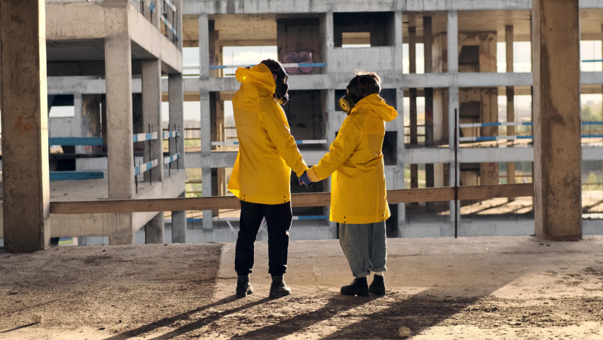 Two stalkers in yellow overalls and gas masks walk through abandoned unfinished building forward towards the sun. back view of two unrecognizable peple in post-apocalypse who hold hands and walk.