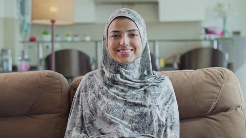 A beautiful modern young Arabic or middle-eastern Muslim woman or female wearing hijab sitting on a couch and looking at the camera confidently and smiling in an interior home setup in daylight