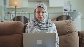 A Modern young beautiful smiling Arabic Muslim or middle eastern woman wearing hijab sitting on a couch and having a conversation over a video call through Laptop in an interior house setup