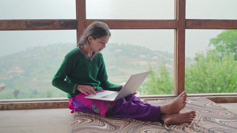 A track in shot of a young girl sitting and studying online with help of the internet and laptop by the window of the house in the remote village region. Distance learning and education concept