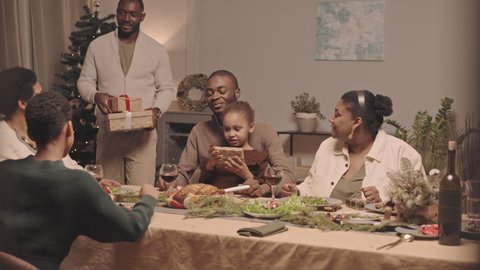 Medium long of young African American man giving Christmas gifts to his mother and brother who sitting at festive table among family, hugging them