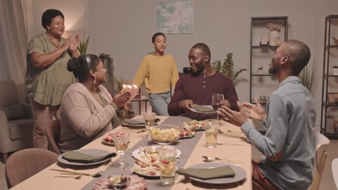 Medium long of two Black girls bringing out and putting birthday cake down on table in front of young happy father, who then blowing out candles and hugging daughters, family members clapping hands