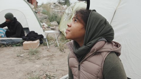 Medium shot of hungry 11-year-old African-American girl in warm clothing sitting in tent at refugee camp, taking tin of food out of bag and looking up