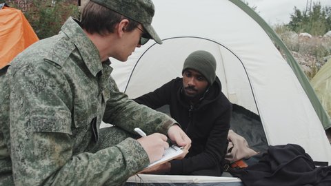 From-above medium shot of young police officer in military uniform taking notes on clipboard while talking to African-American refugee man sitting inside his tent at immigrants camp