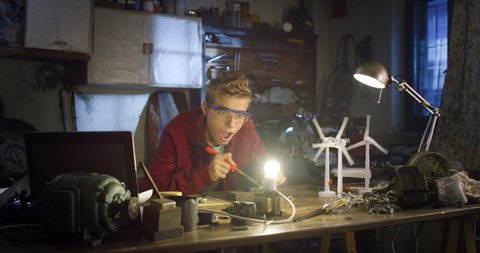 Cinematic shot of curious teenage boy with specific glasses is working on engineering project of sustainable green energy for school in his father workshop.